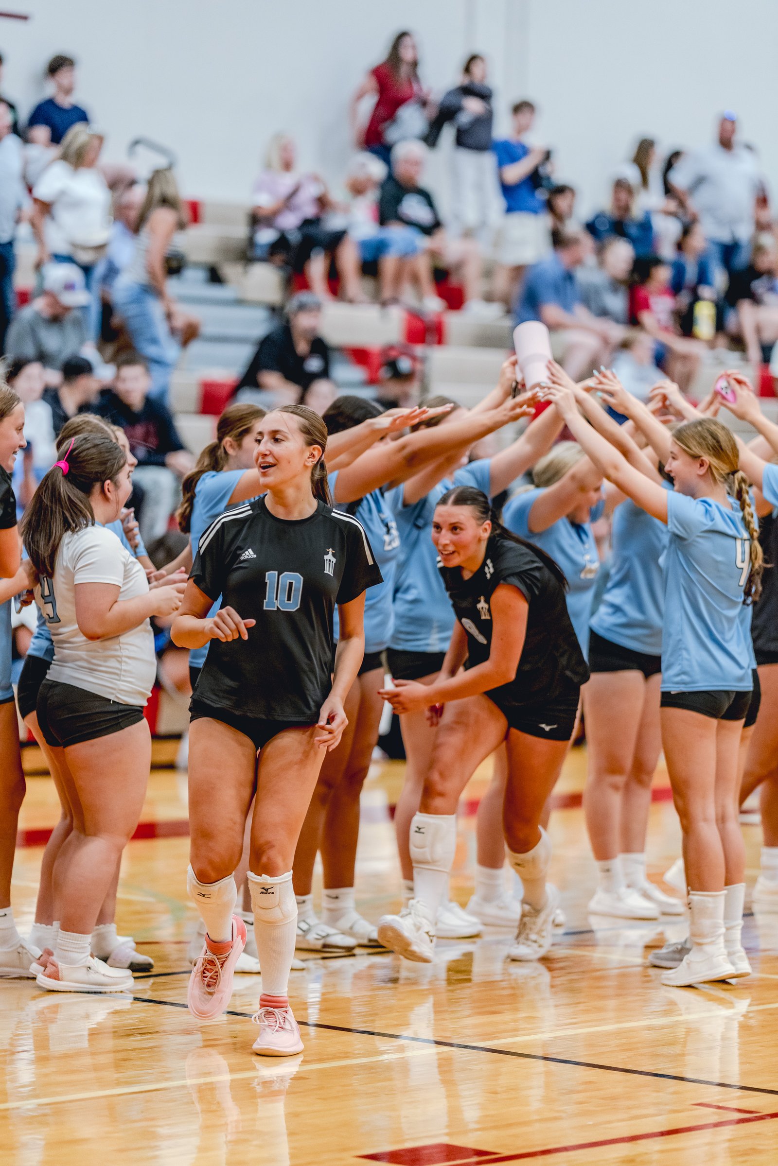 Players in black and light blue jerseys celebrate on the court, with one player in black wearing jersey number 10.