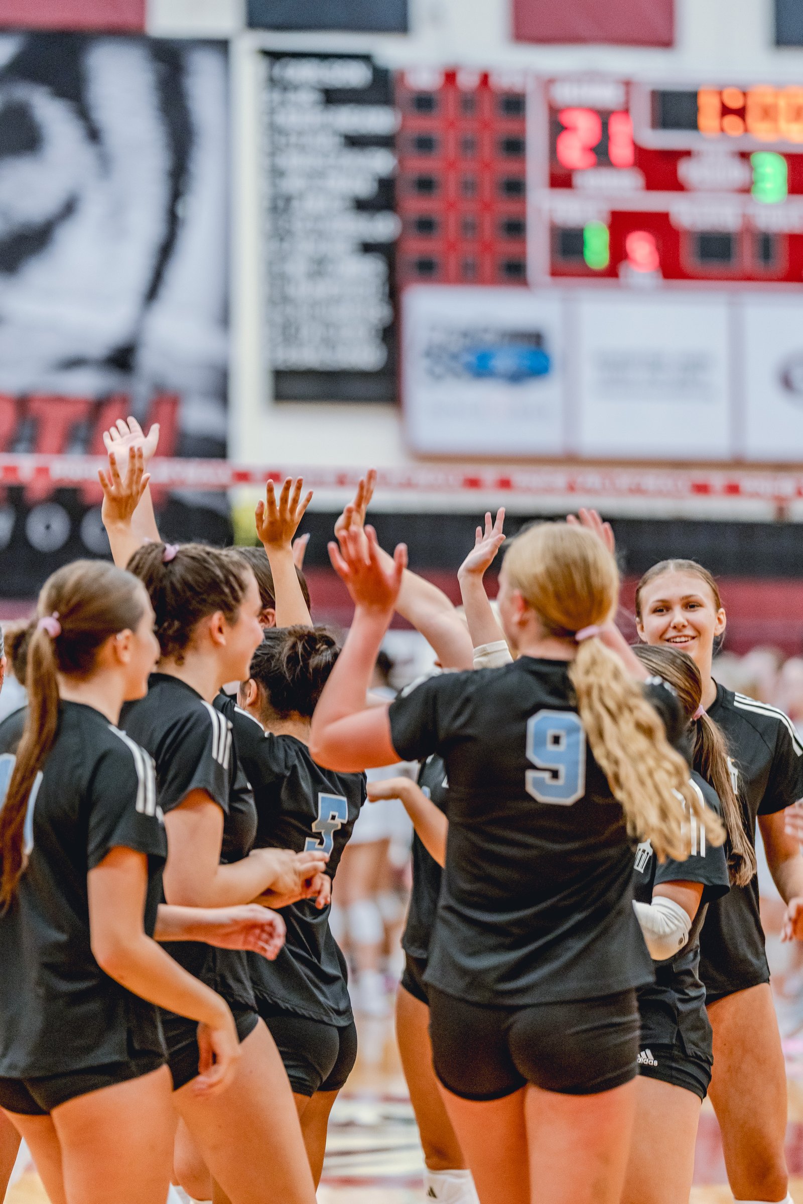 Players in black jerseys with light blue numbers celebrate on the court, with a scoreboard visible in the background.