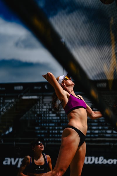 A player in a purple top and black bottoms reaches up with her right arm extended.