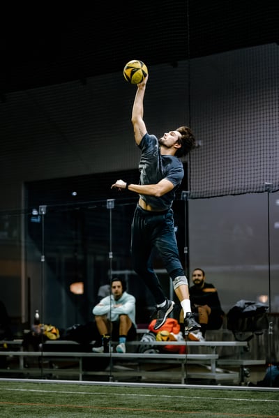 A player in a gray shirt jumps with one arm extended overhead for the ball.