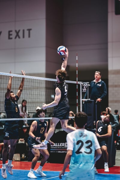 A player in black jumps and reaches overhead for the ball while a player in navy reaches up at the net.