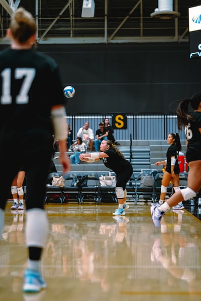 A player in black crouches low with arms extended toward the volleyball.