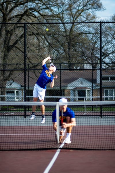 A player in blue jumps and swings a racket at the ball while another player in blue crouches low.