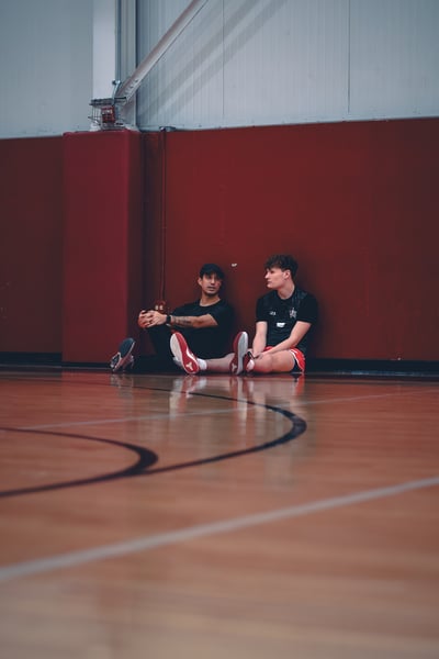 A player in black and a player in black shorts sit on a basketball court leaning against a red wall.