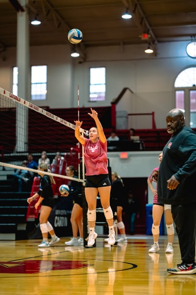 A player in a pink shirt and black shorts jumps with arms extended toward the ball.