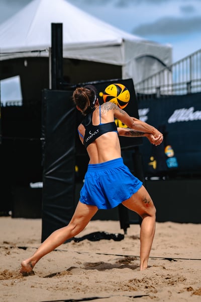 A player in blue shorts and a black top extends her arms to hit the ball on a beach court.