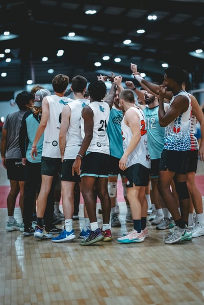 Players in white and blue uniforms huddle with arms raised in a gym.