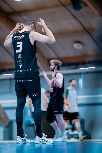 A player in black adjusts orange straps over his head while a player in black and red gestures with open hands.