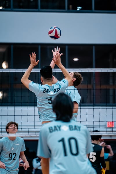 A player in a light blue shirt reaches up for the ball while another player in a light blue shirt jumps near the net.