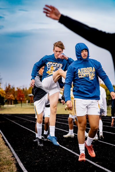 A player in a blue hoodie kicks his leg up while other players in blue hoodies and white shorts stand on a track.