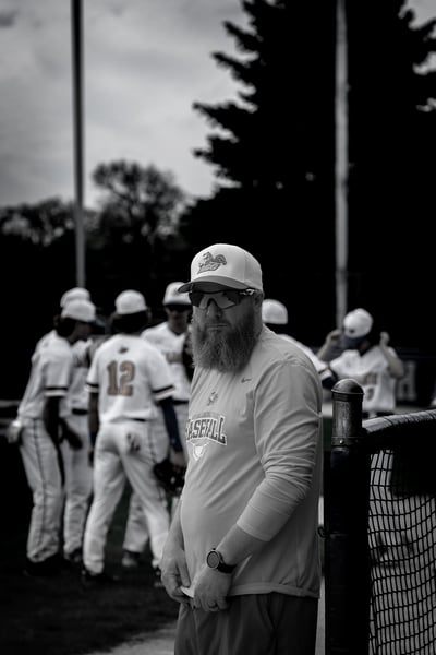 A man in a light-colored shirt and cap stands with his hands clasped, while players in white uniforms stand behind him.
