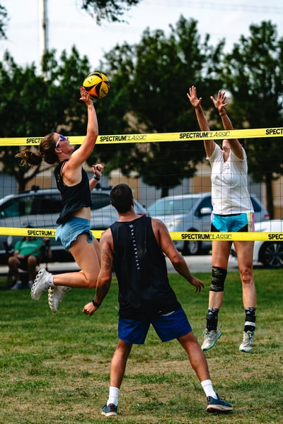 A player in black jumps to hit the ball while a player in white reaches up and a player in black watches.