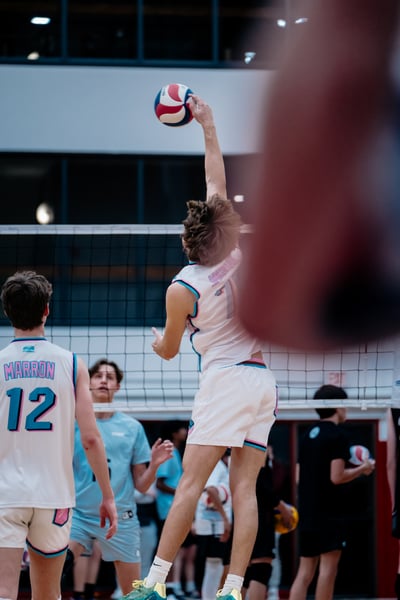 A player in white reaches up with the ball, while a player in white with blue trim looks on.