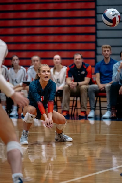 A player in a dark blue uniform with orange sleeves crouches low, hands open, as a volleyball flies overhead.