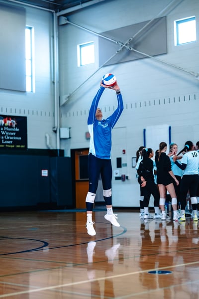 A player in a blue top jumps with arms extended overhead for the ball.