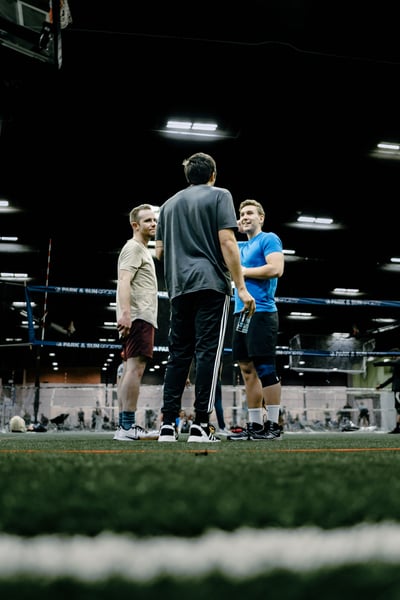 A player in blue stands with arms crossed while talking to players in tan and gray.