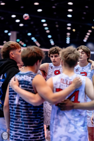 Players in white and blue jerseys huddle with arms around each other, a volleyball is in the air.