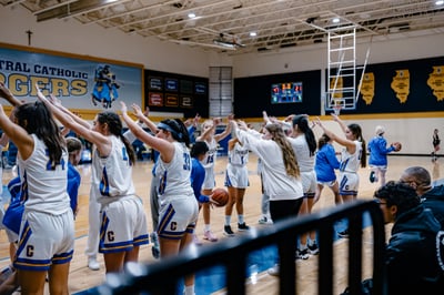 A player in white and blue holds a basketball while players in white and blue raise their hands.