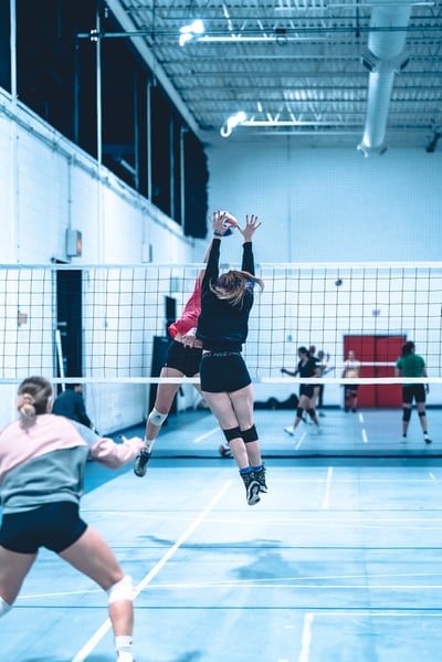 A player in black reaches overhead for the ball while a player in red jumps next to her.