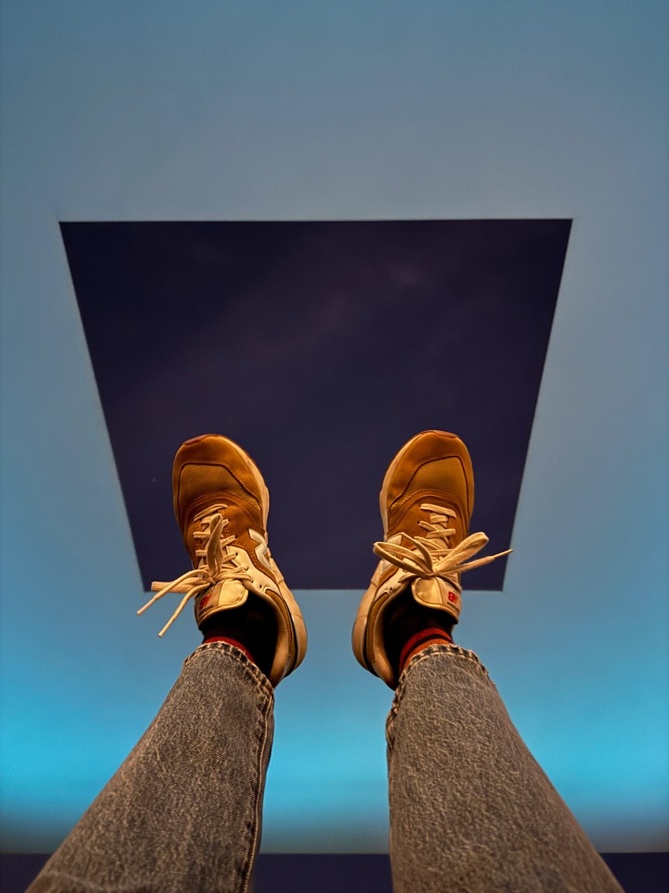 Photo of James Turrell's Skyspace exhibition. The sky is visible in a rectangular viewport with light blue-ish light around the viewport. The photographer has her feet up in the air so you see them in the viewport. She's wearing New Balance sneaks.
