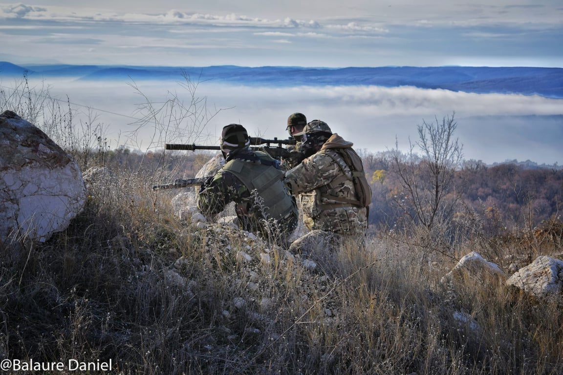 Prim-plan al unui lunetist Airsoft DTS în costum ghillie ce își ia poziția în spatele unui adăpost