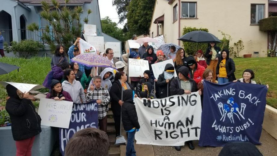 People hold signs in the rain to support a group of unhoused and housing-insecure families who have occupied vacant homes in the northeast LA neighborhood of El Sereno.
