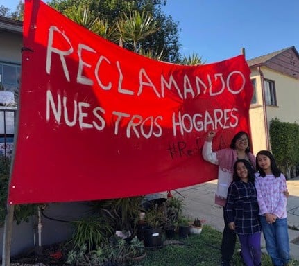 Martha Escudero raises her fist and poses with her two daughters in front of a banner with "Reclaiming Our Homes" written in Spanish.