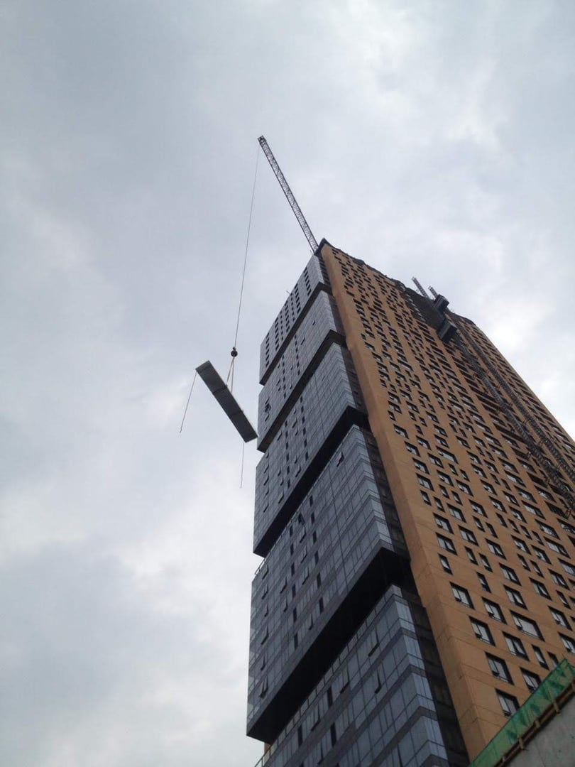 Crane lifting a beam next to a tall, modern building against a cloudy sky.