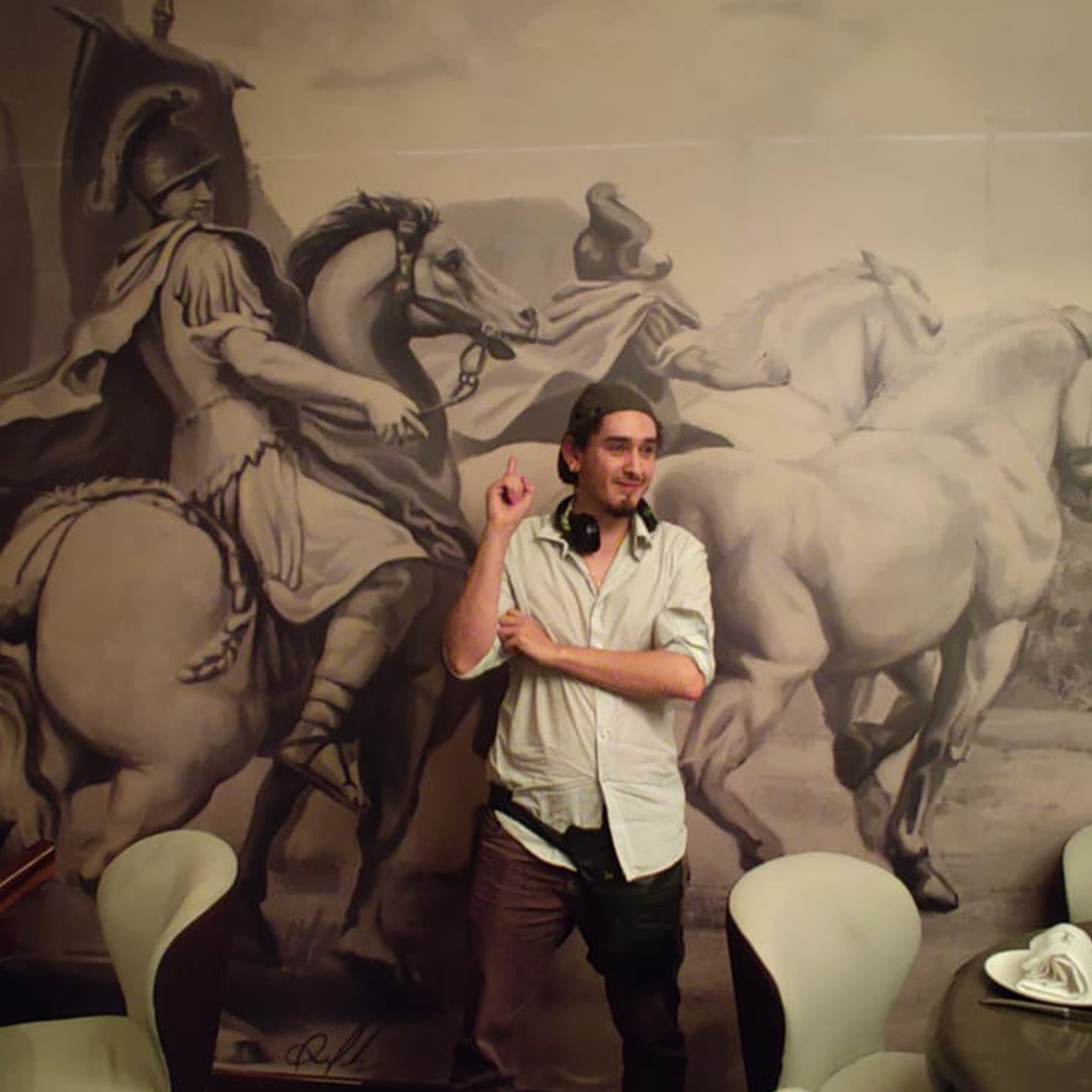 Man in casual attire poses in front of a large mural depicting a historical scene with horses and a soldier on horseback.
