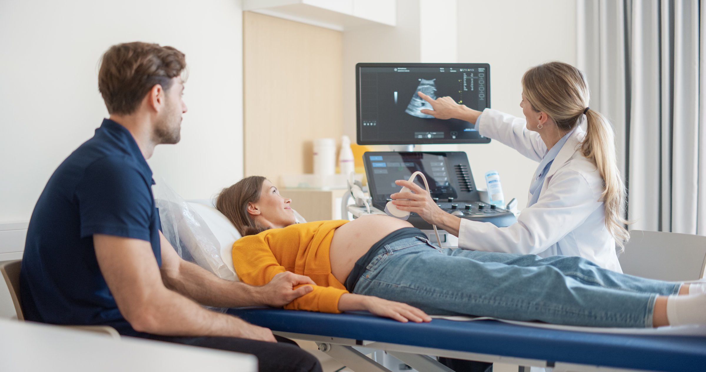 Modern Prenatal Ultrasound Session in a Hospital. Female Doctor Examining a Fetus on Computer Monitor. Pregnant Woman Smiles as Her Boyfriend Offers Comfort Beside Her in the Medical Office