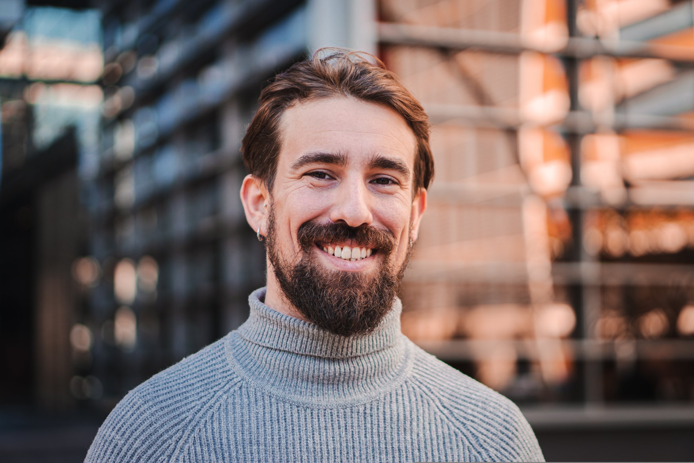 Close up portrait of awesome young caucasian male smiling and looking at camera at the background city office buildings. High quality photo