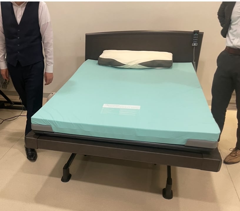 Adjustable bed with turquoise sheet and pillow, flanked by two men standing on either side in formal attire.