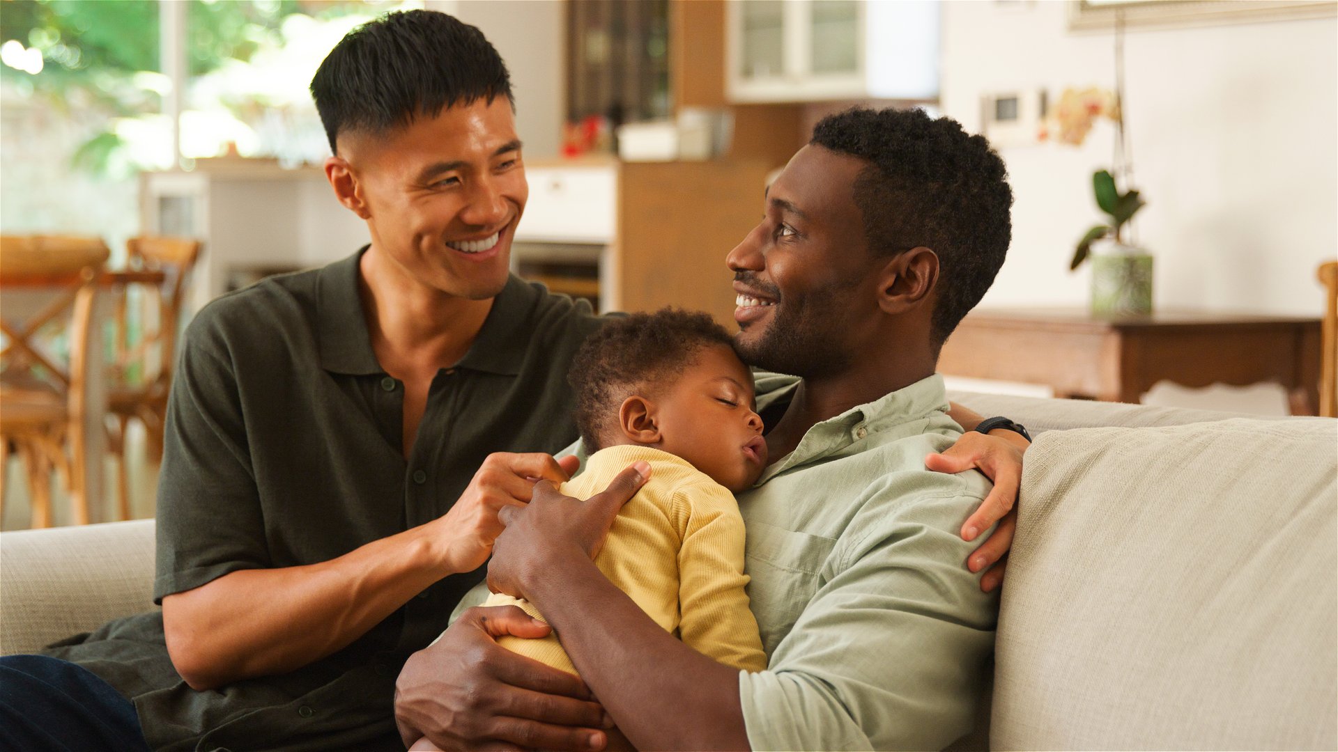 Same sex male couple joyfully interact in a cozy living room with a sleeping child.