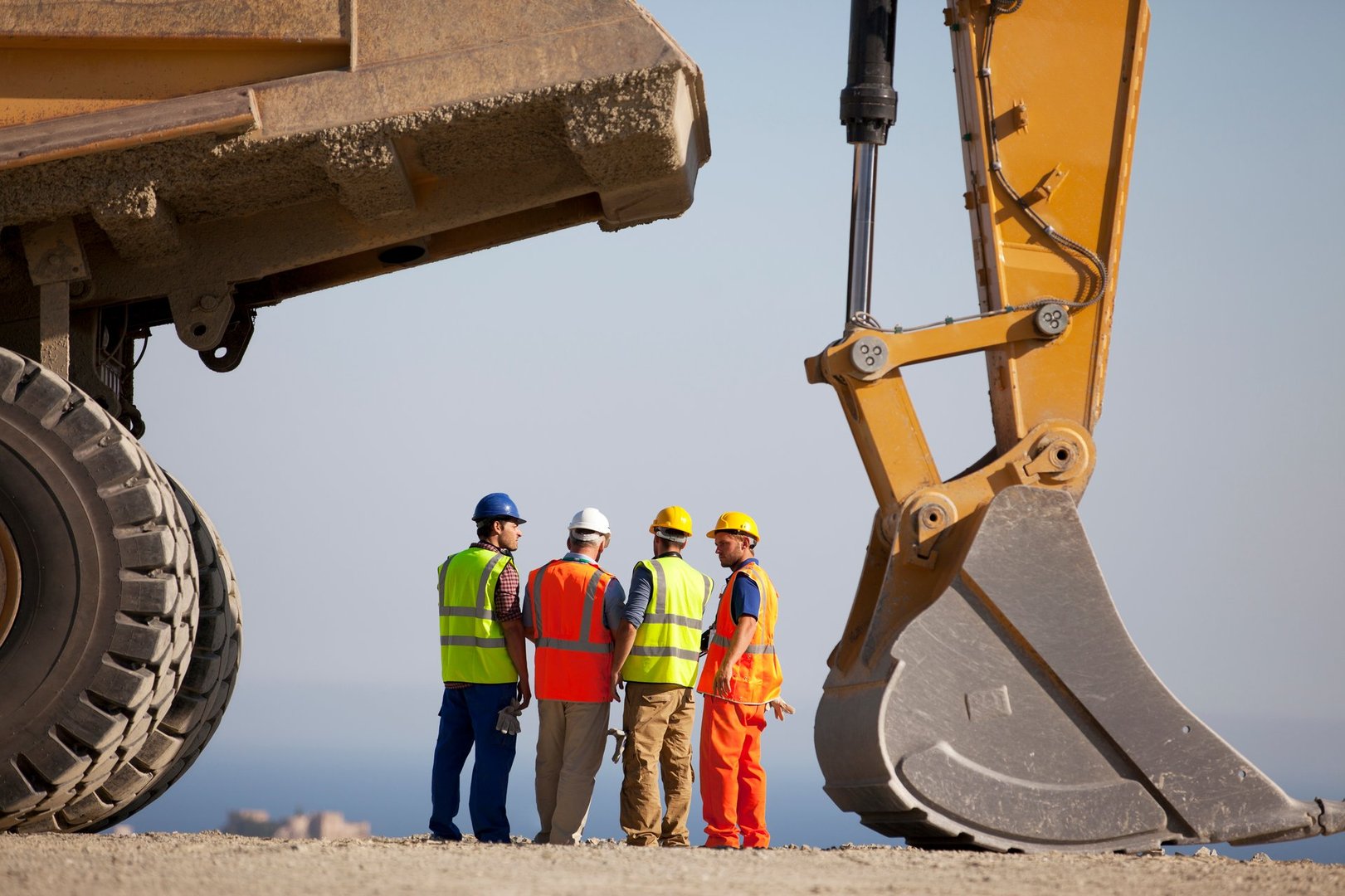 Four construction workers in safety gear stand near heavy machinery on a construction site, discussing plans.