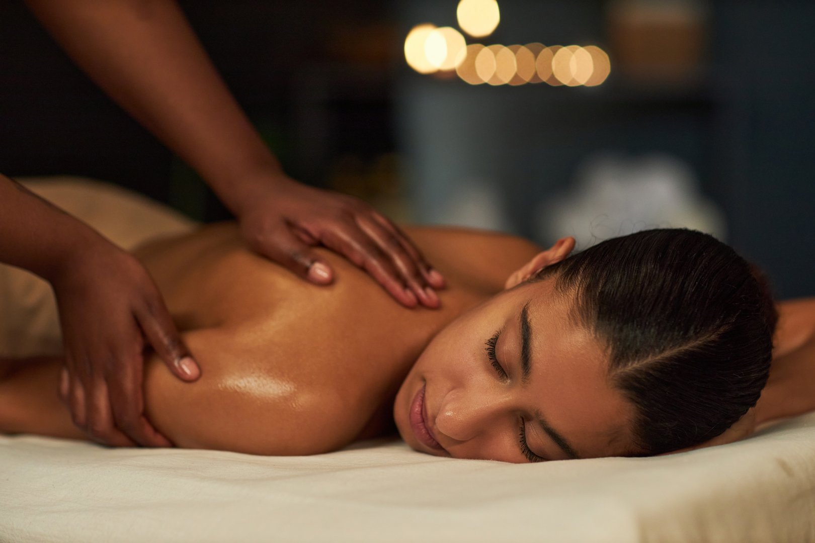 Woman relaxing while receiving massage in spa setting with candles creating soothing ambiance, hands of massage therapist gently working on shoulders and back