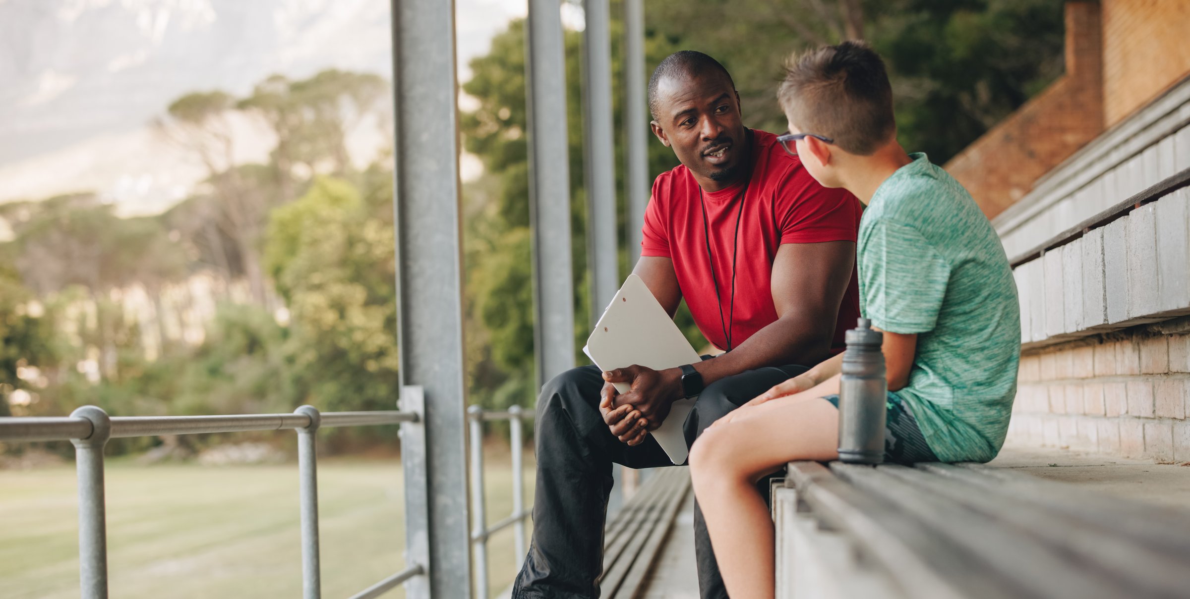 School coach talking a young student while sitting outside. Physical education teacher motivating a young boy in elementary school. Child mentor giving support and guidance.