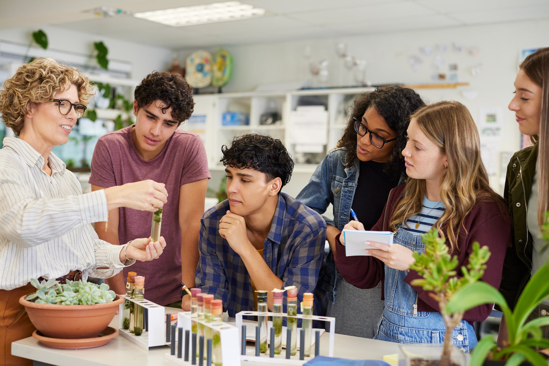 Mature science teacher showing sample in test tube to a group of multiethnic students during classroom activity. High school guys and girls gathered around table to observe biology experiment involving plant specimen. Group of young college classmates working on science project at school.