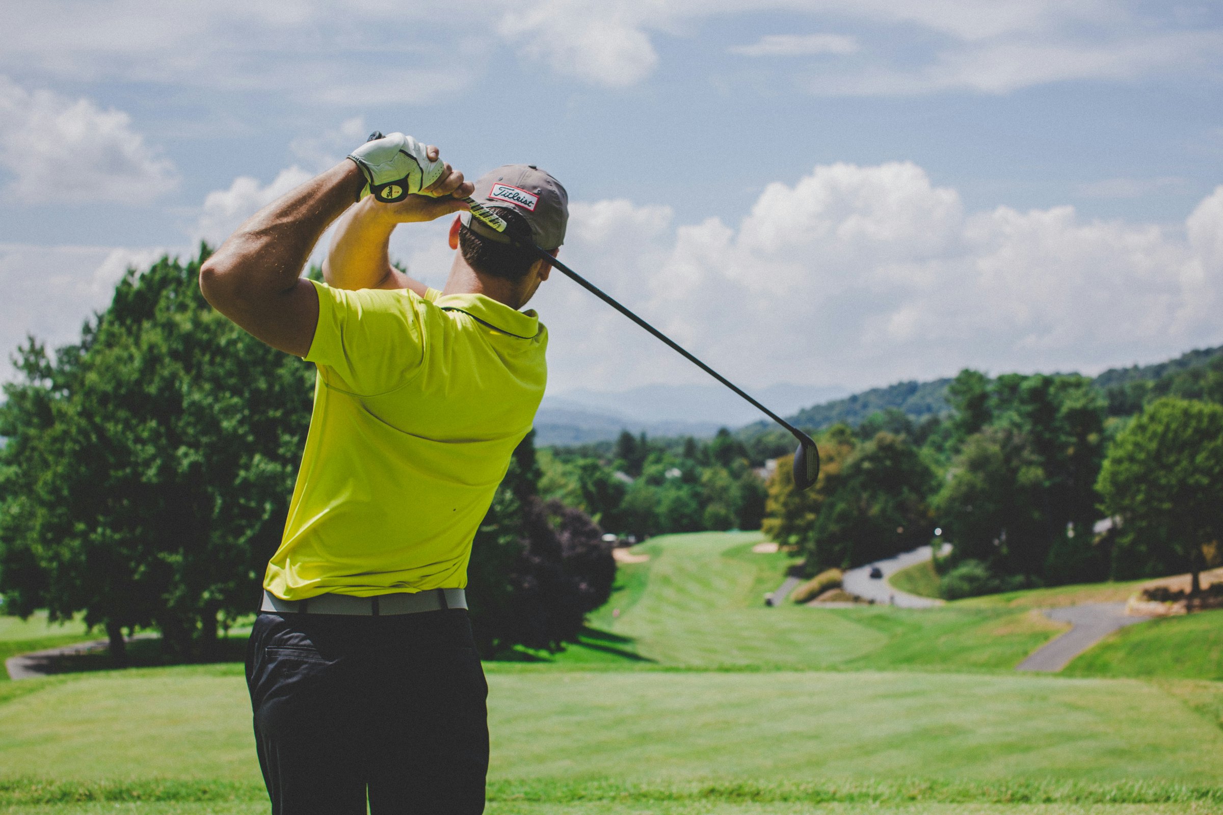 Golfer in yellow shirt swings club on a sunny day, overlooking a green course with trees and distant hills.
