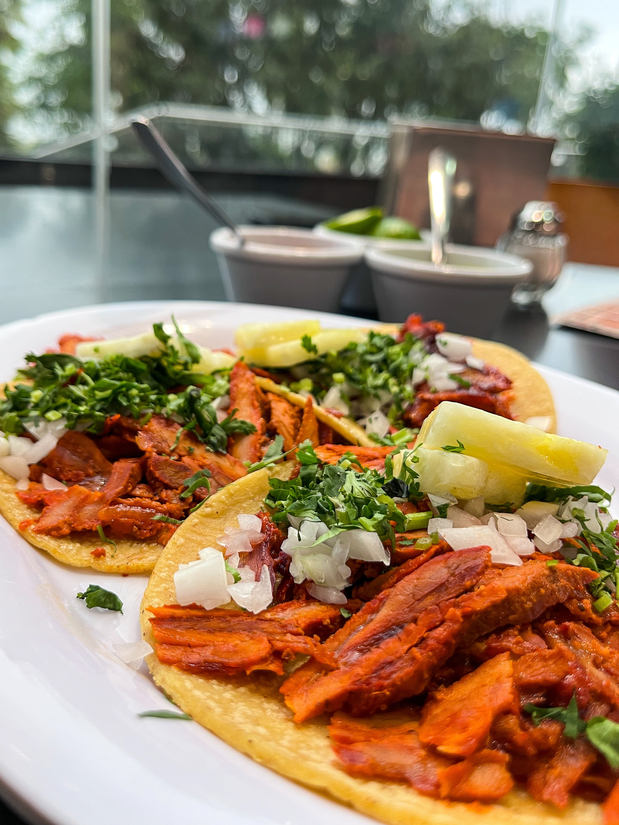 Close-up of two tacos al pastor on corn tortillas, garnished with fresh pineapple, cilantro, and onion. The dish is served at a taquería in Mexico, with salsa bowls, lime, and condiments visible in the background.