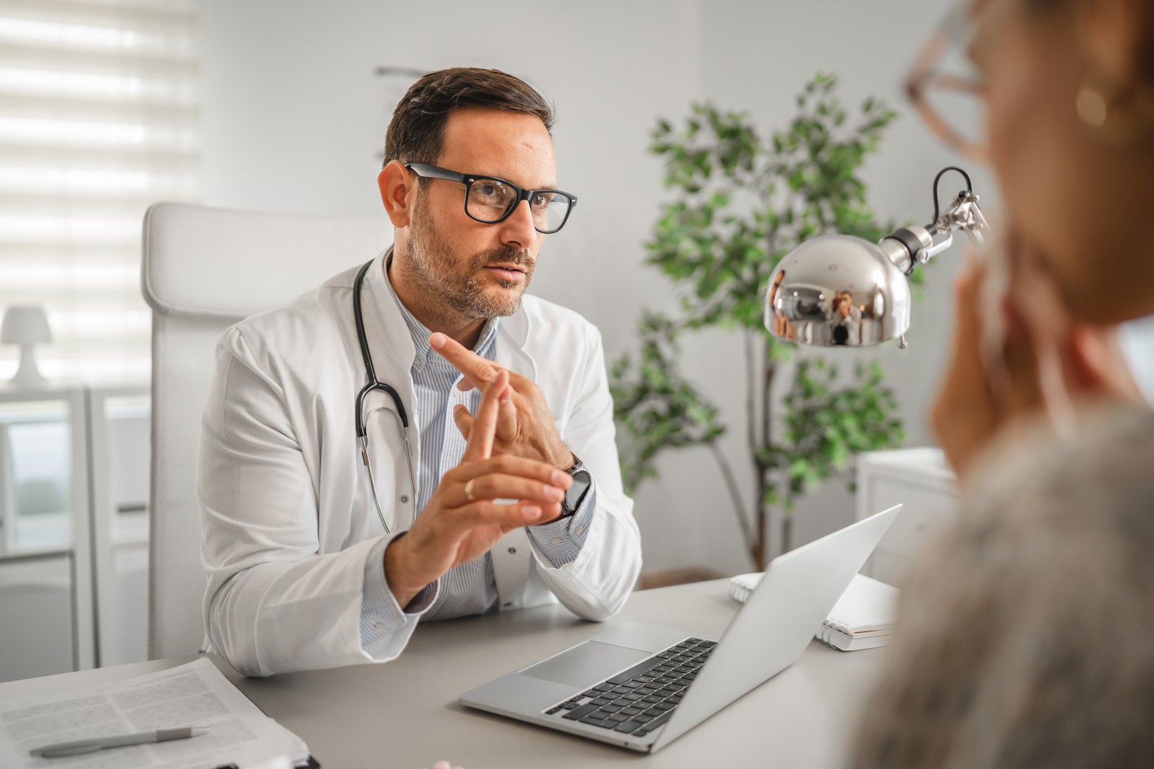 Male doctor wearing a lab coat and stethoscope, sitting at a desk and explaining diagnosis with hand gestures to a female patient during a healthcare consultation in an office