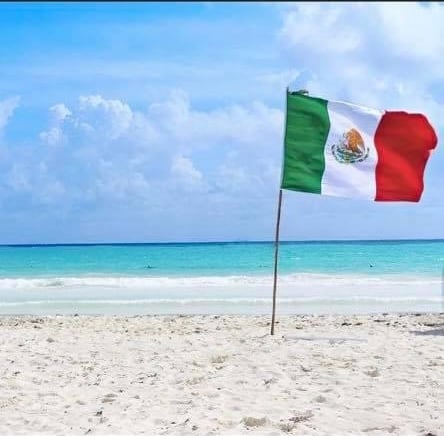 Mexican flag on a sandy beach with turquoise water and cloudy sky in the background.