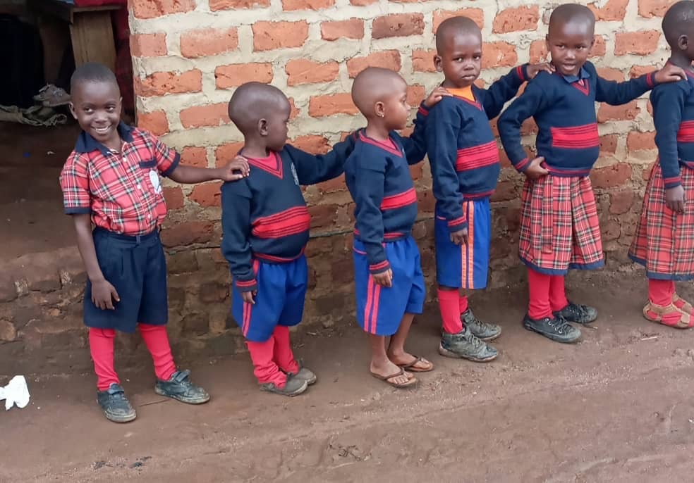Children in school uniforms stand in a line against a brick wall, some smiling and others looking forward.