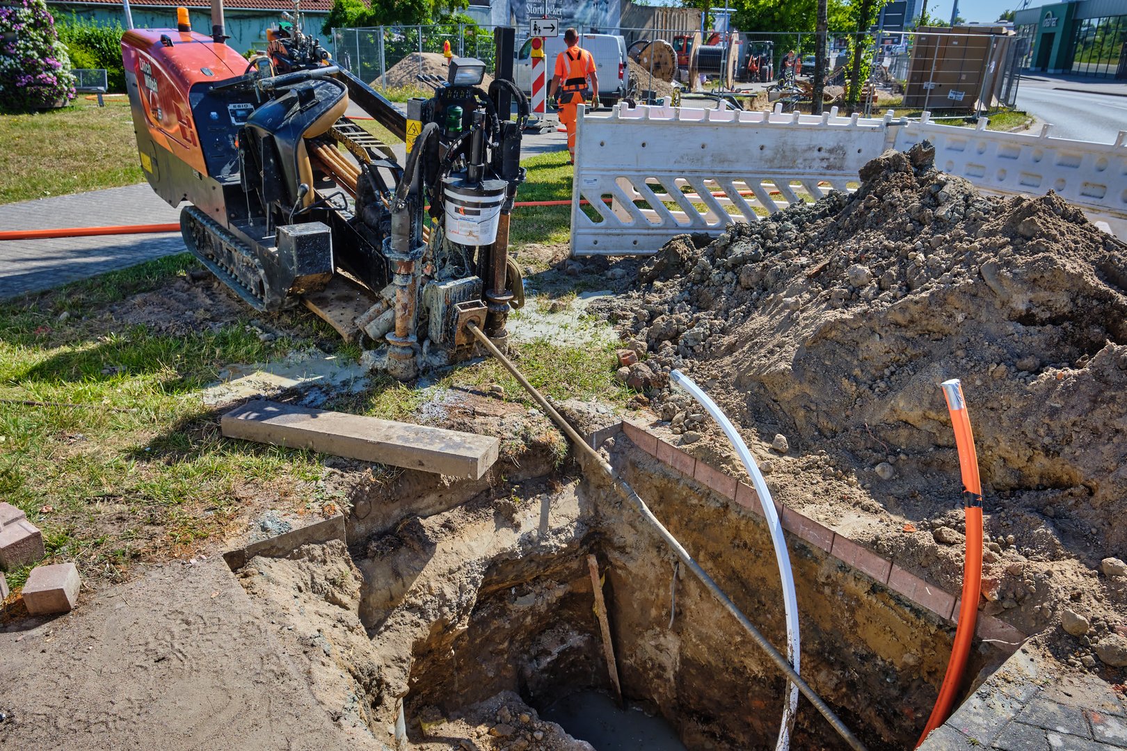 July 1, 2025 - Stralsund-Germany: Small horizontal drilling machine with drill by start pit at roundabout corner, orange and white cables, sand heaps, fences and distant roadworks.