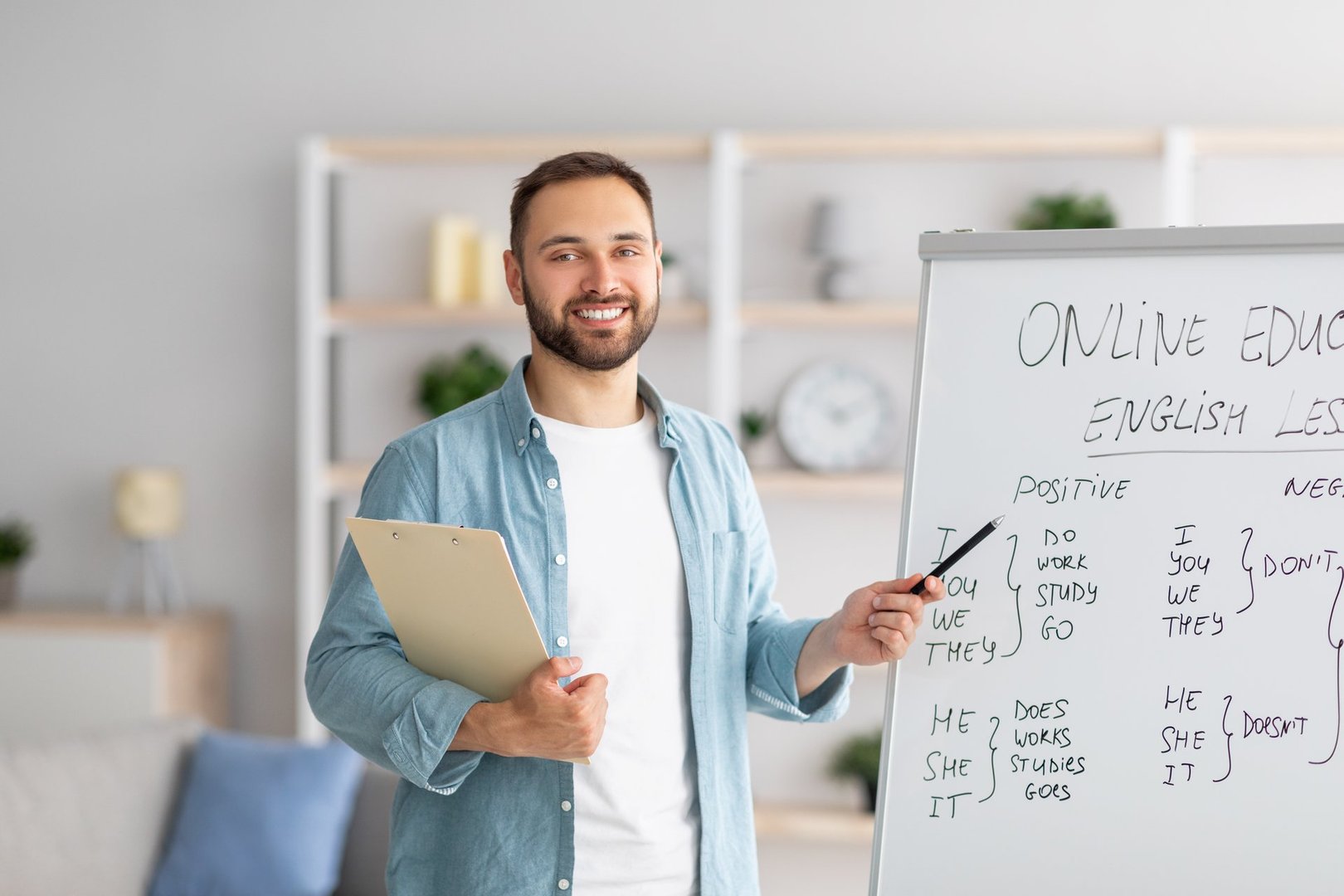 Cheerful young male teacher pointing at blackboard, explaining grammar, giving online English lesson from home office. Distance education and web tutoring during coronavirus lockdown
