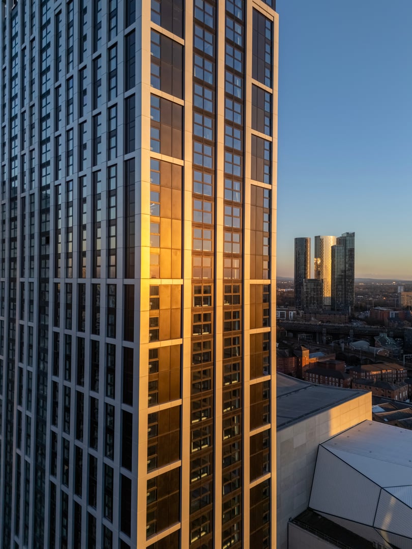 This photograph has not been created by AI other than to adjust tone and contrast. An aerial photograph of a skyscraper in downtown Salford, Greater Manchester, Northwest England. The image shows a closeup of a skyscraper bathed in the golden rays of the sun and was created on a beautiful February afternoon as the sun was setting, The wider Manchester skyline can be seen in the distance, bathed in the last of the day's sunshine.
