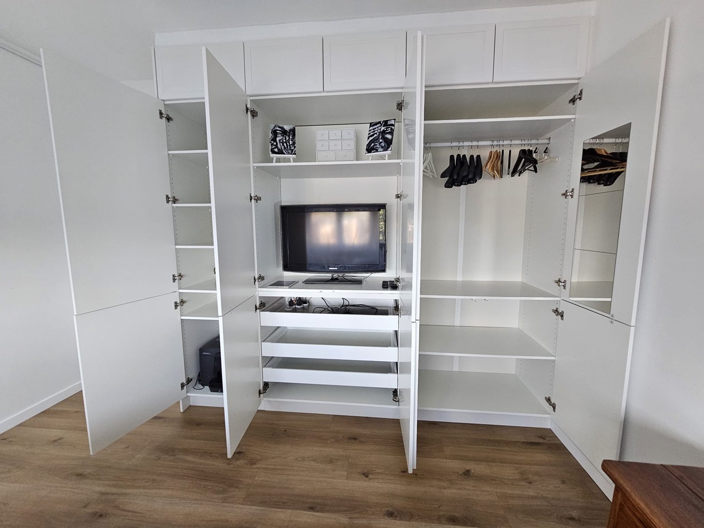 Open white closet with a TV, shelves, drawers, and hanging clothes on a wooden floor.