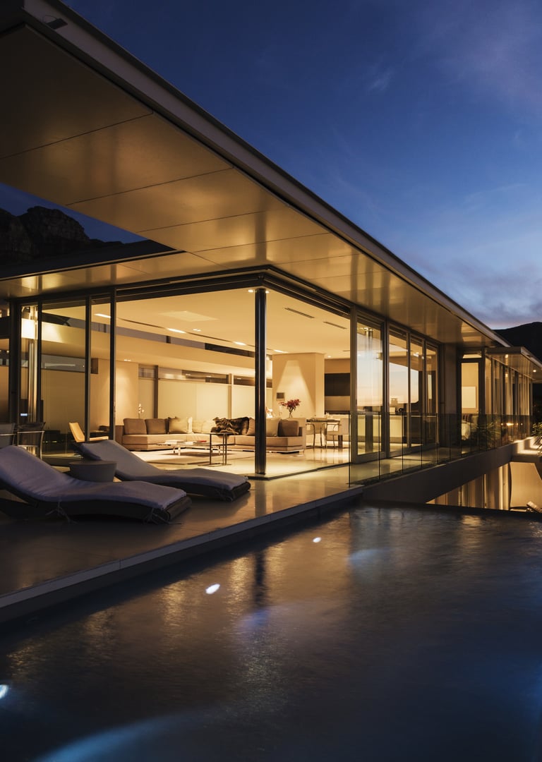 Modern glass-walled house at dusk with illuminated interior, outdoor lounge chairs, and a reflective infinity pool.