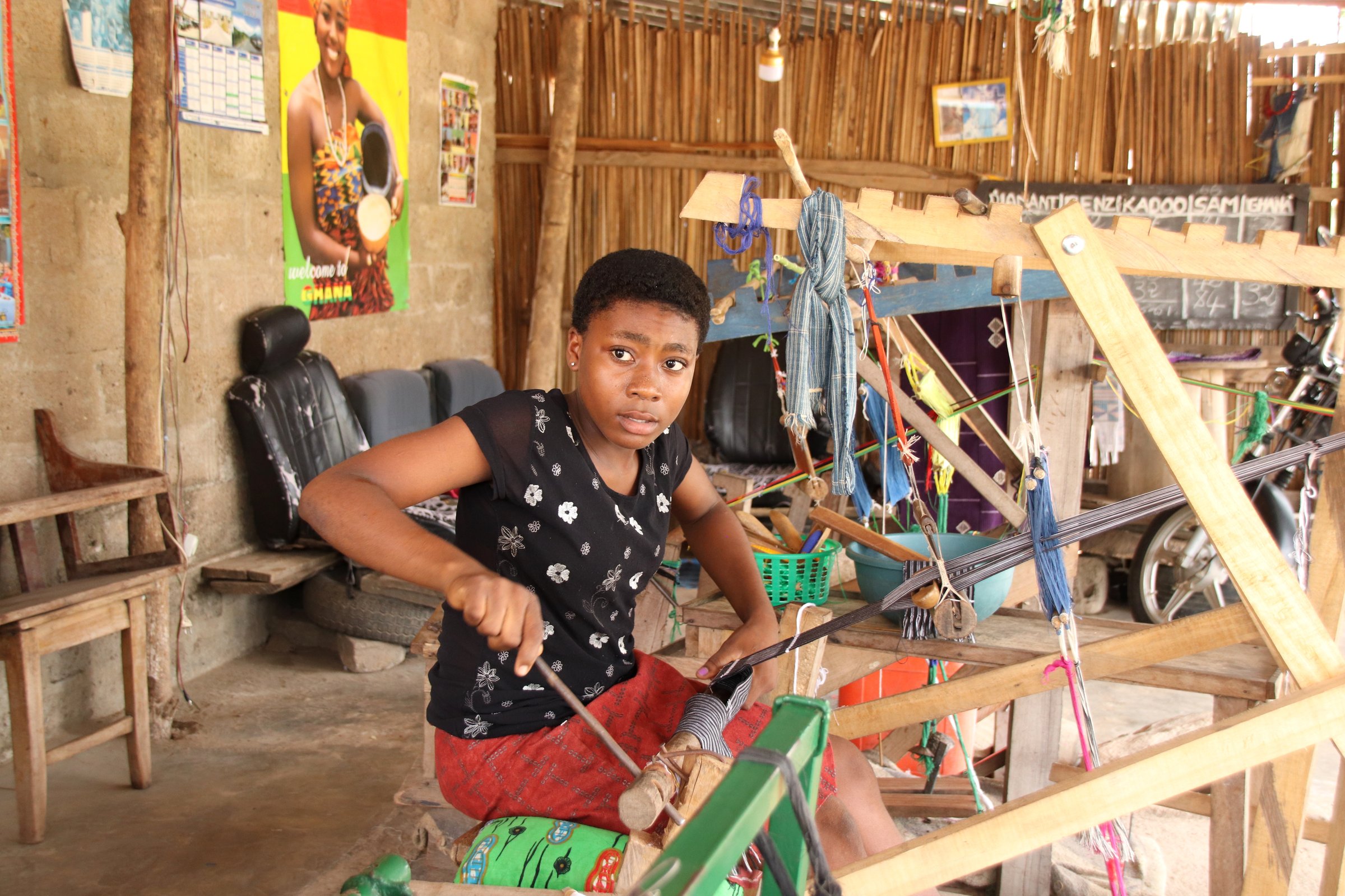 Kpalimé, Togo - June 27, 2019: Young woman of a weaver family weaves traditional Kente fabrics on the side of the road in Kpalimé, Togo, West Africa. They use traditional weaver looms.