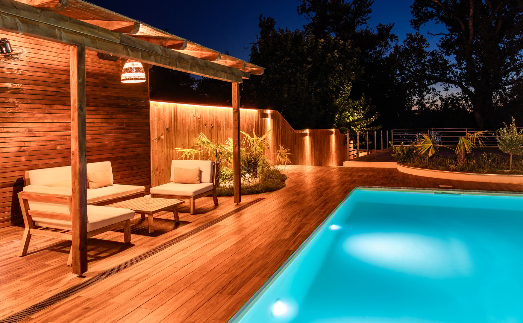 Modern outdoor lounge area with wooden pergola and swimming pool at night. Luxury backyard illuminated with soft lighting