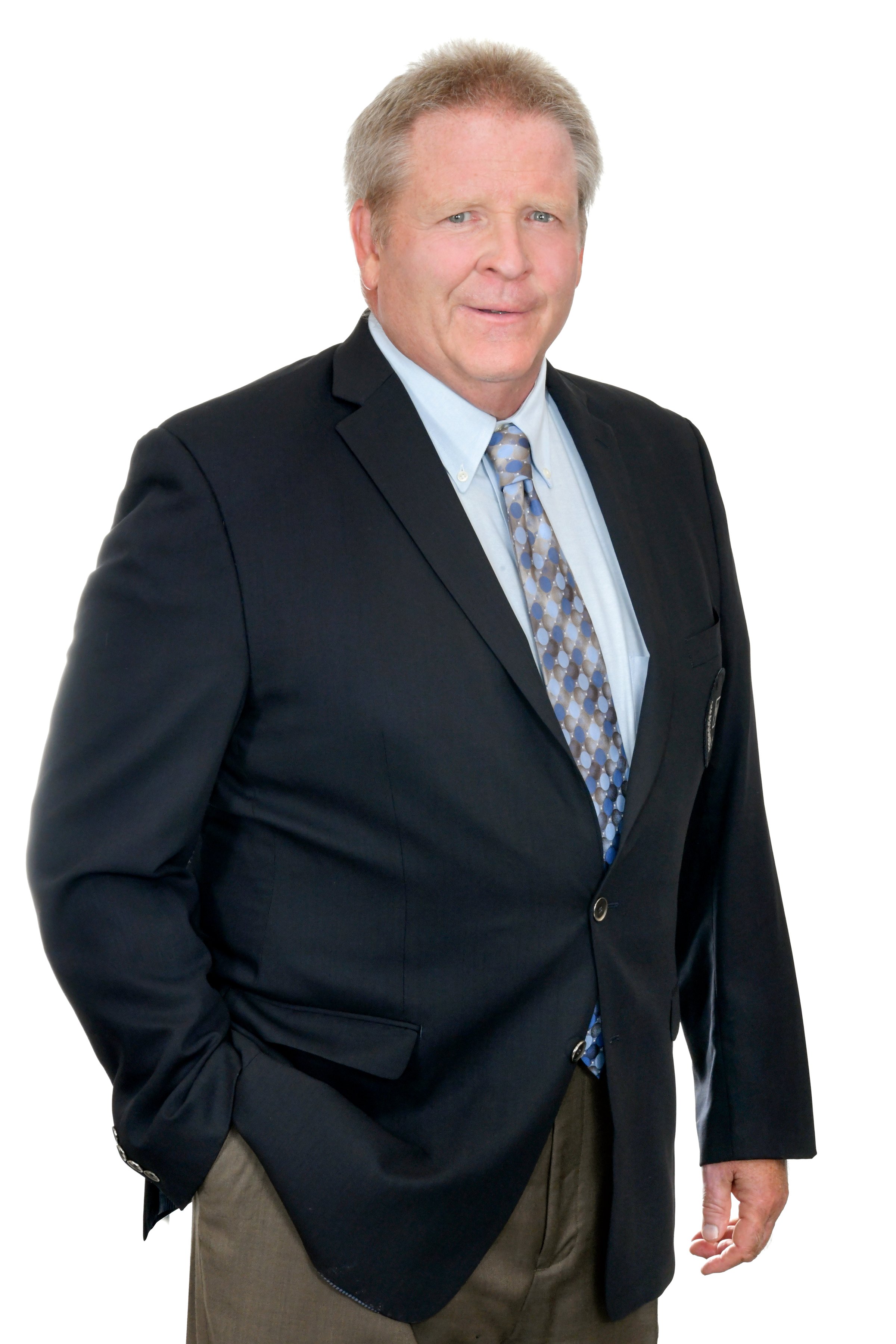 Man in a suit and tie with a light blue shirt, posing against a white background, hand in pocket, smiling slightly.